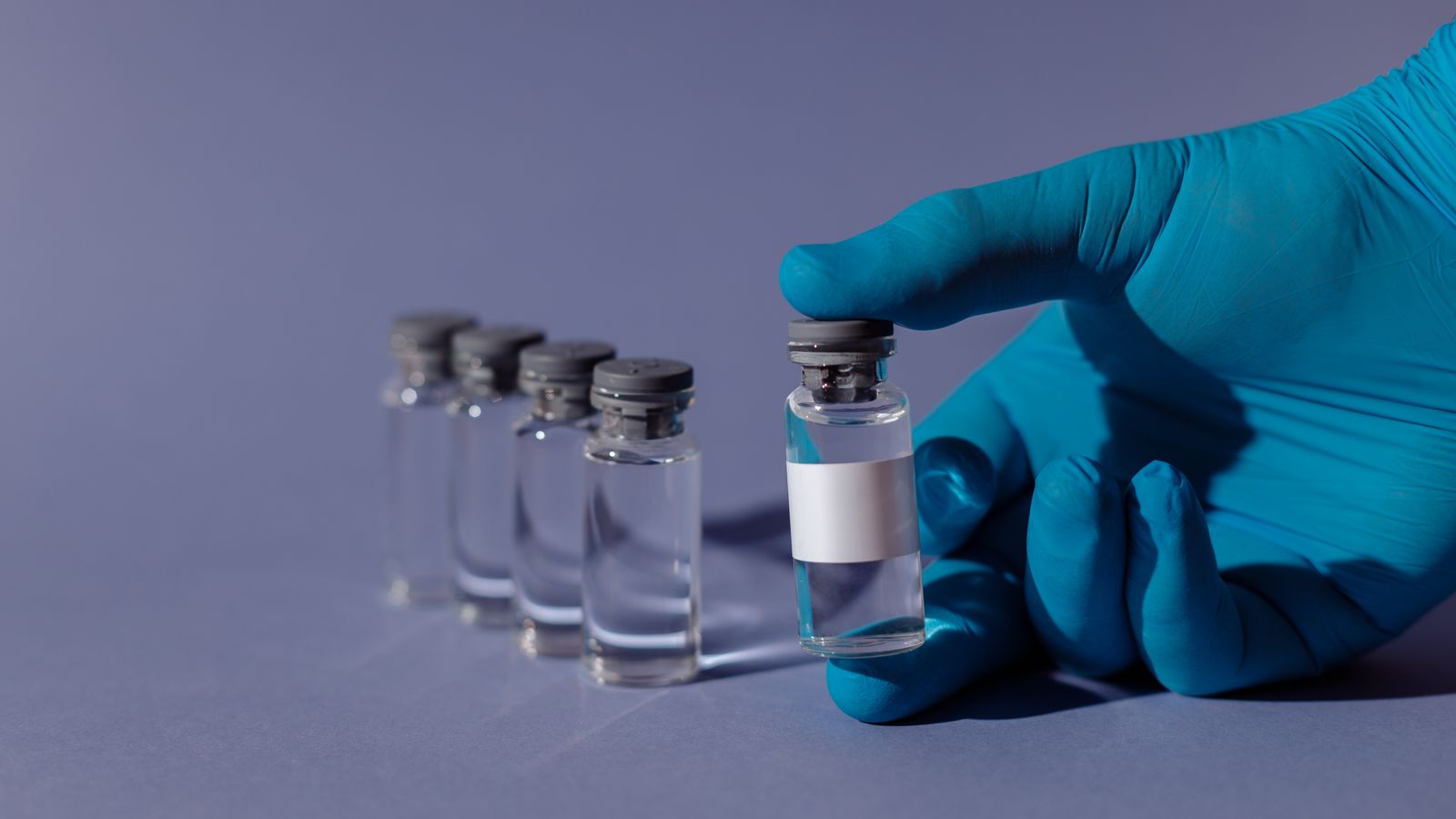 A blue-gloved hand holding a transparent vial filled with liquid against a clinical background.
